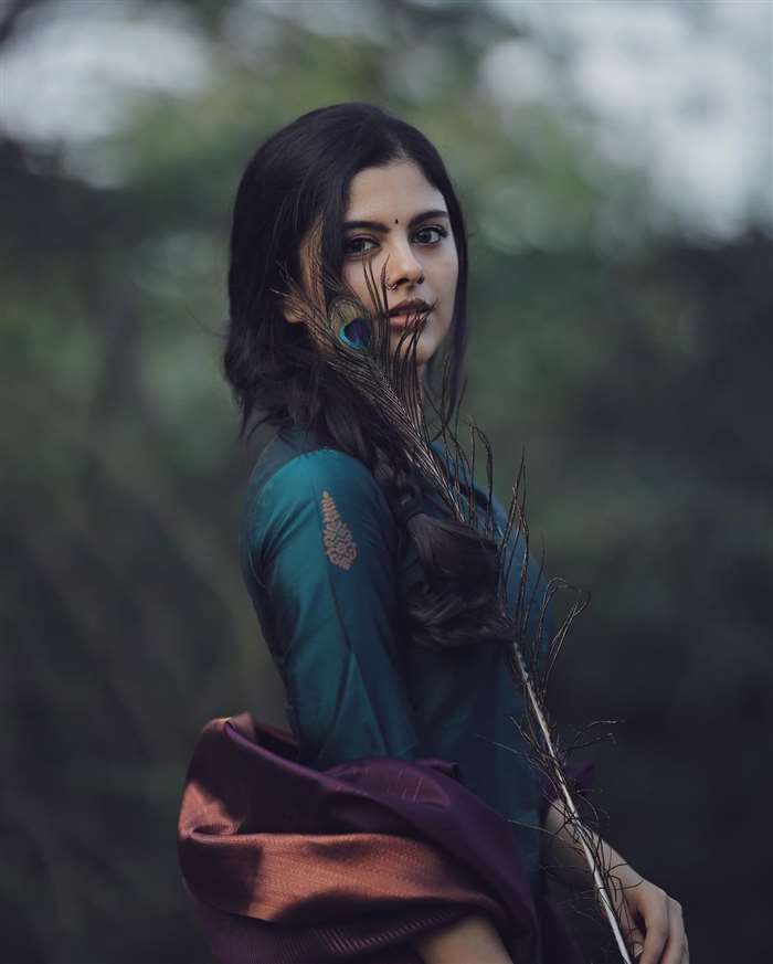 Sanjana K is captured in a portrait wearing a white, long-sleeved blouse and draped with a patterned fabric in a soft, warm setting.