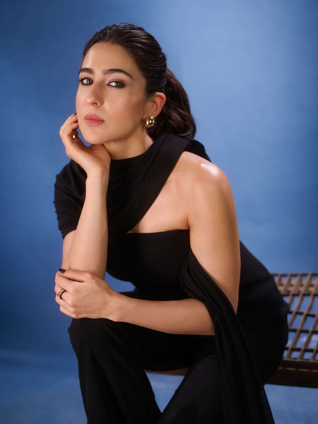 Sara Ali Khan posing in a black draped outfit against a vibrant blue studio background, holding black fabric.