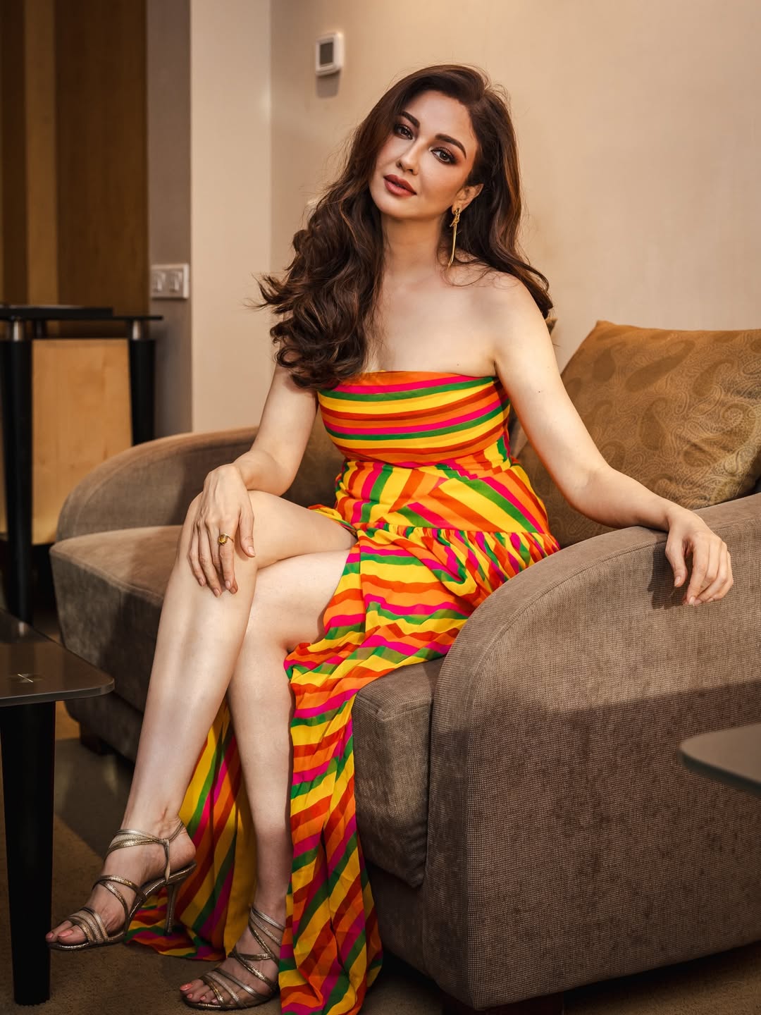 Saumya Tandon is seated on a large, upholstered sofa wearing a brightly colored, flowing garment and strappy sandals.
