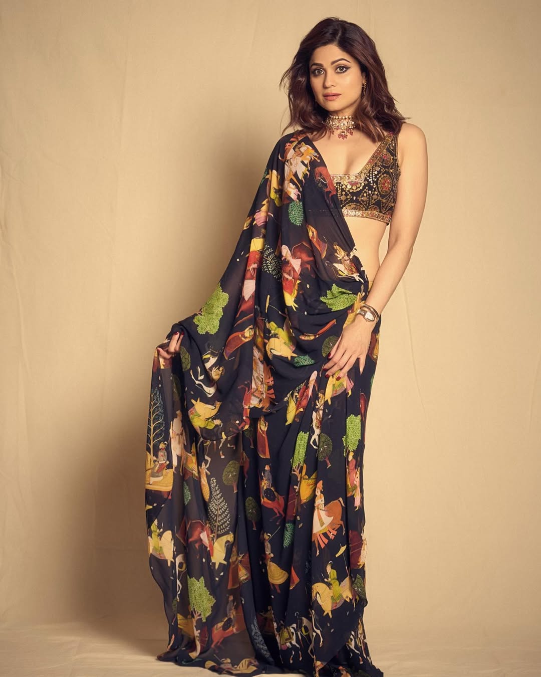 Shamita Shetty stands centrally, wearing a coordinated ensemble in peach, pink, and green, set against a backdrop of metallic hanging objects.