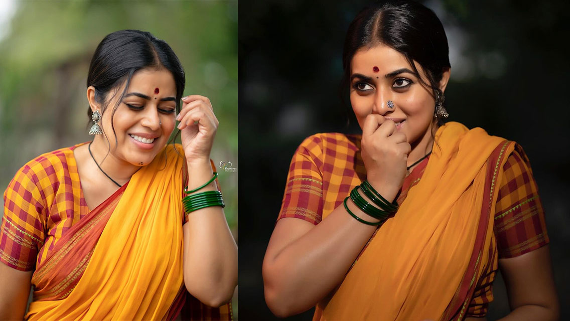 Shamna Kasim is draped in a bright yellow saree with red and maroon borders, wearing green bangles and a red bindi, posing outdoors.