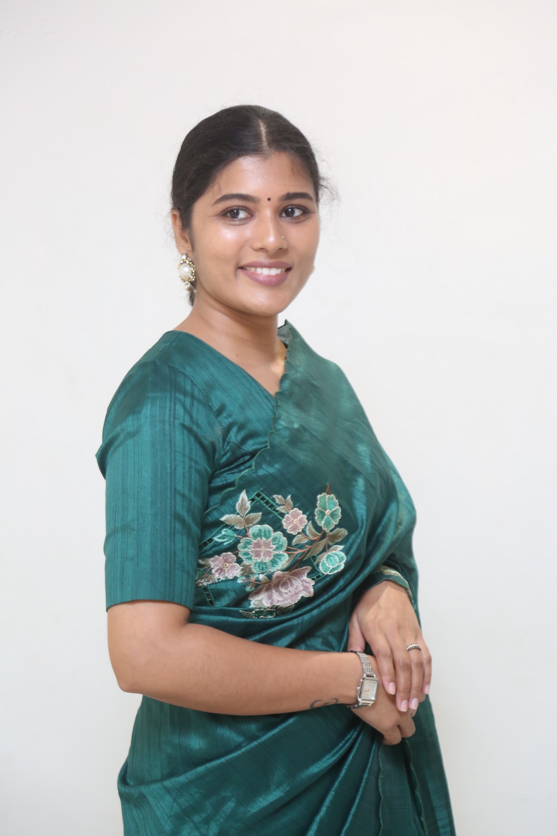 Sharanya Pradeep poses elegantly against a plain background, draped in an exquisite deep teal saree.