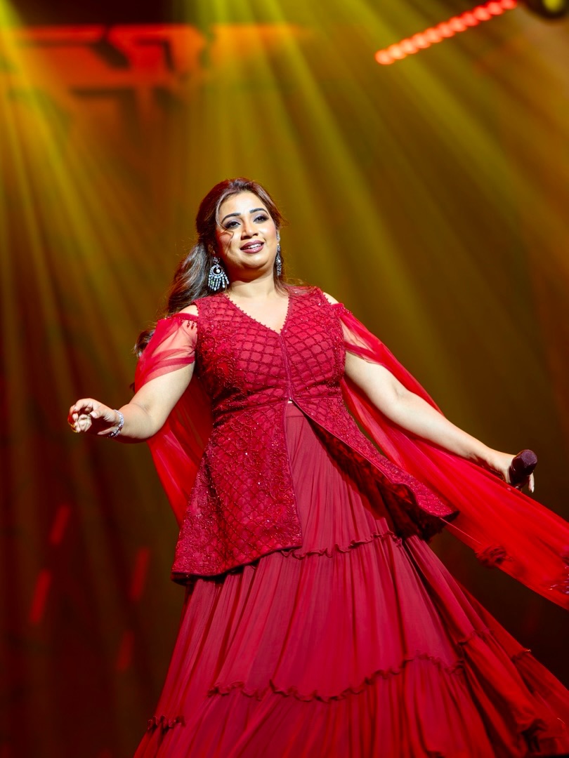 Shreya Ghoshal in an elaborate, light-colored, heavily embellished traditional outfit with flowing silver detailing and soft waves.