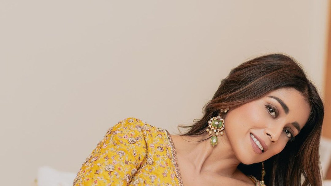 Shriya Saran reclining in a vibrant golden yellow traditional outfit, smiling warmly for the camera.
