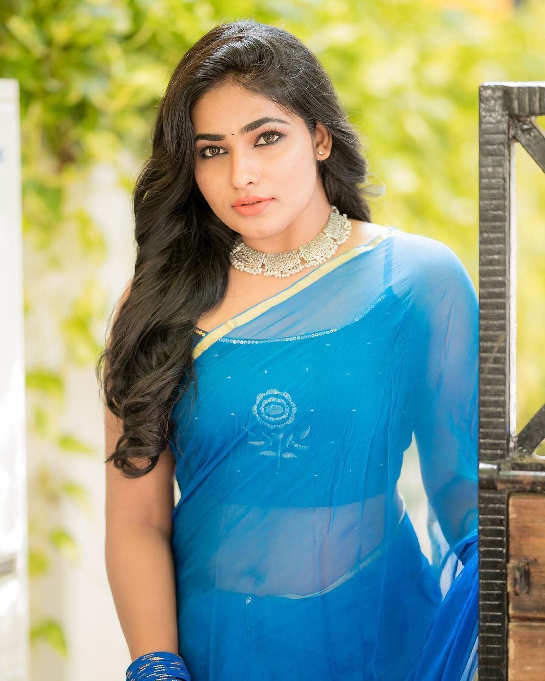 Shruthi Selvam stands near a concrete ledge wearing vibrant red fabric and a blue skirt, looking at the camera.