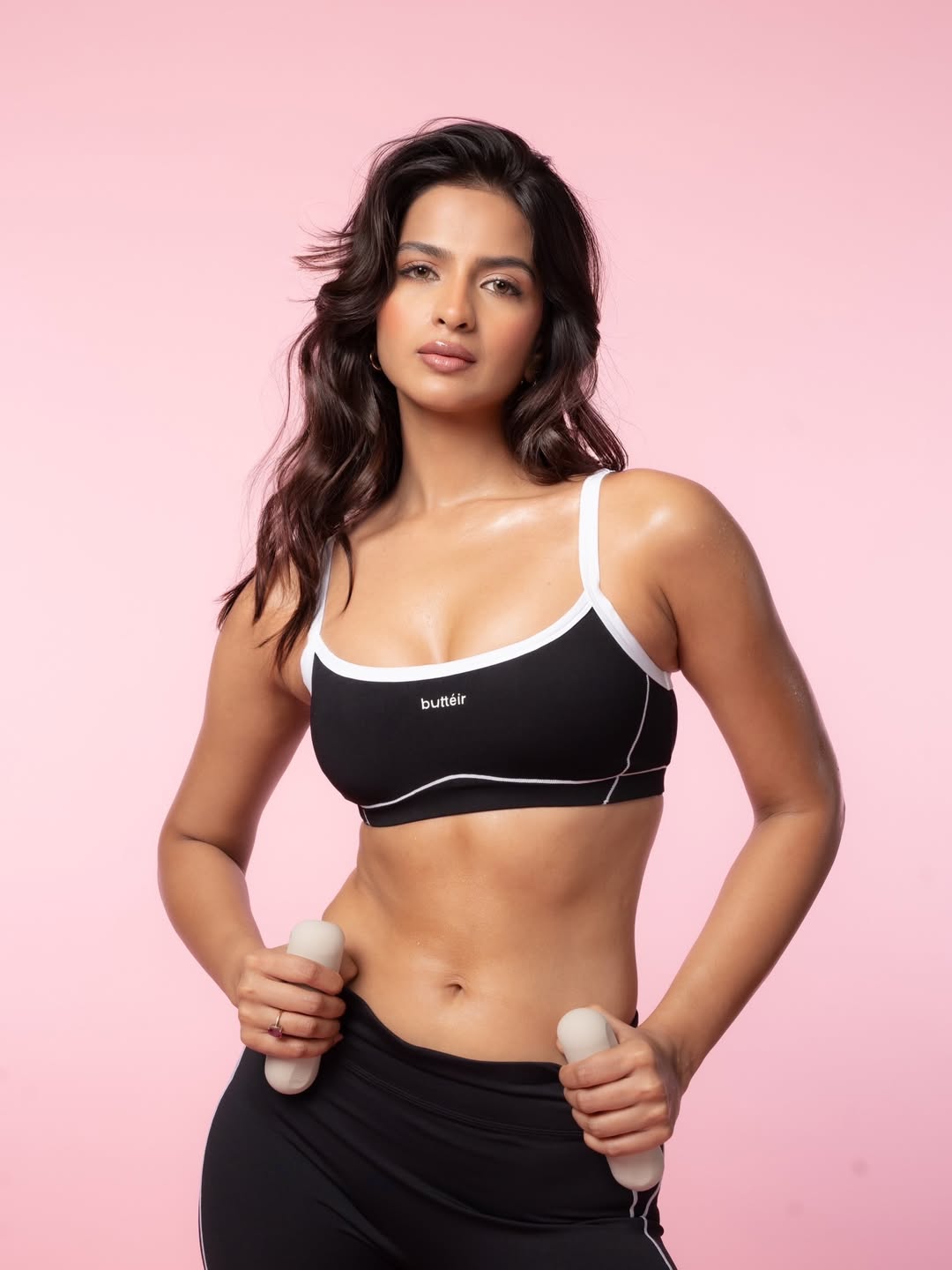 Shruti Chauhan posing in athletic wear against a solid pink background, wearing a black crop top and matching leggings.