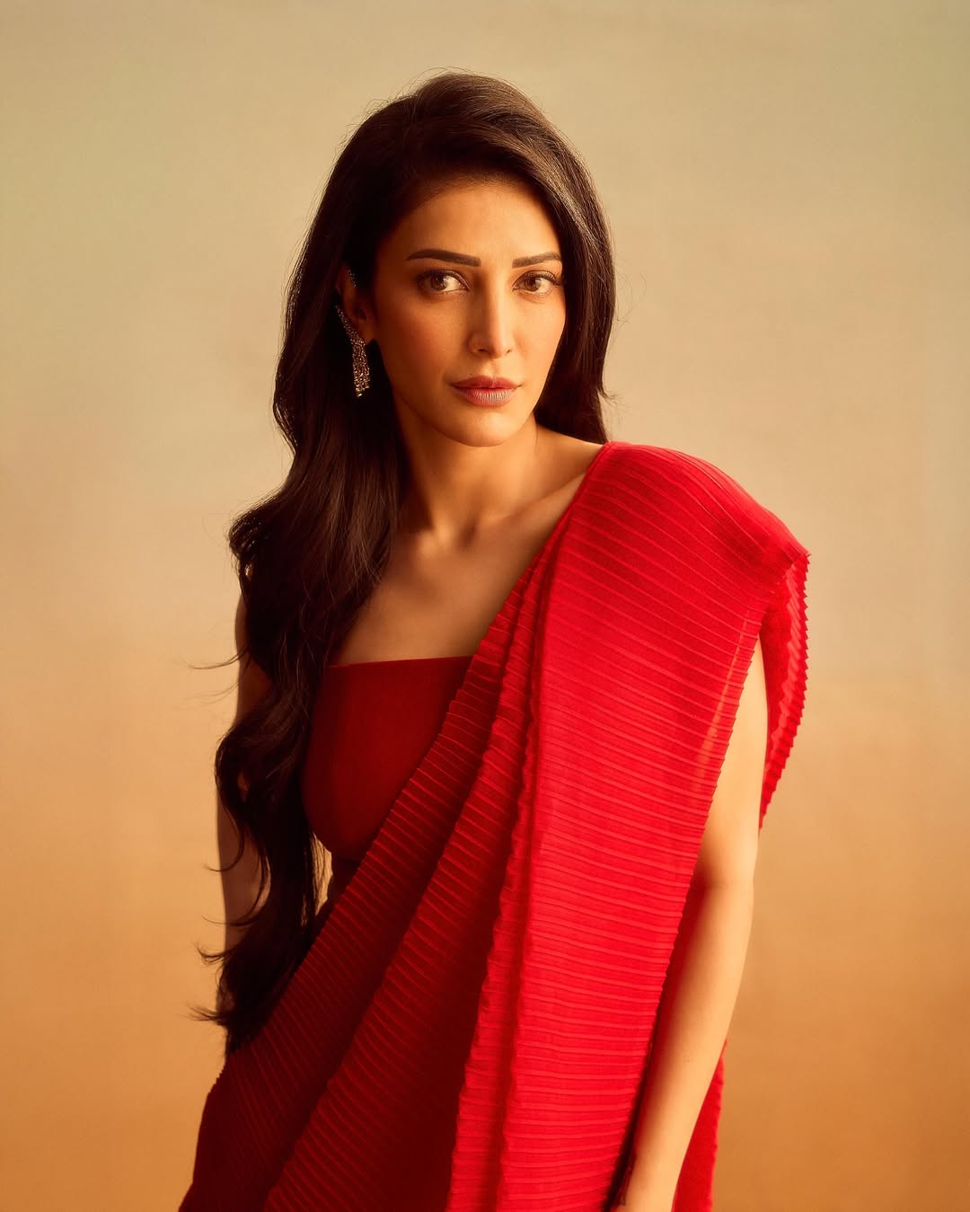 Shruti Haasan standing in a full-length photograph wearing a vibrant, flowing deep red garment against a light beige background.