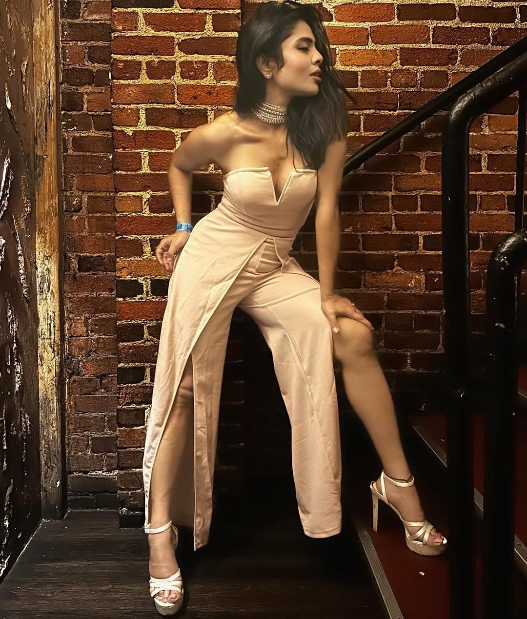 Simi Nair poses gracefully in a shimmering, light gold gown against a dark, featureless background, highlighting her elegant posture.