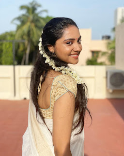Smruthi Venkat is draped in light fabric and adorned with white floral garlands in a lush outdoor setting.