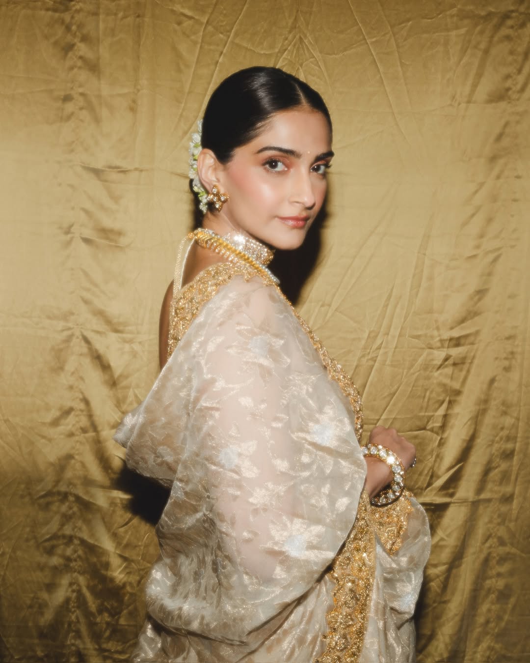 Sonam Kapoor poses indoors wearing a royal blue top, patterned shawl, and lime green trousers with gold detailing.
