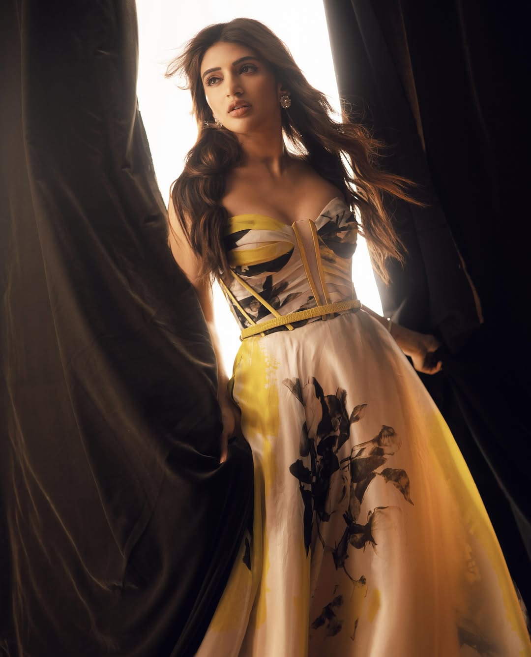 Sreeleela stands framed by dark drapery, wearing a light-colored gown with dark patterns and golden accents, with dramatic lighting.