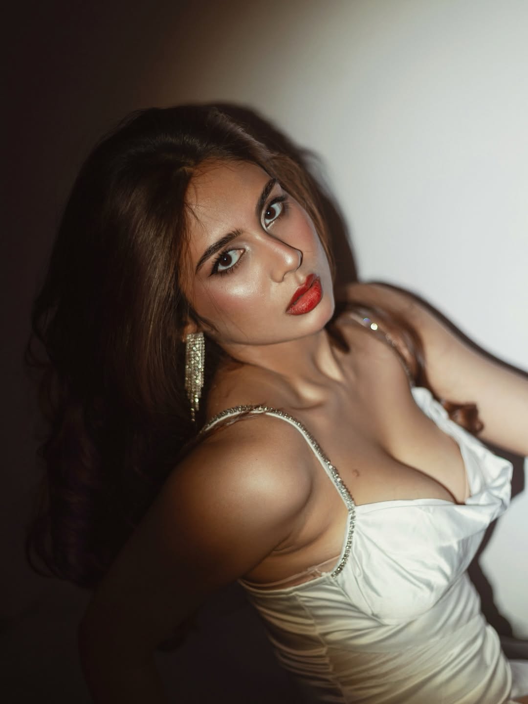 Srushti Bannatti posing elegantly in a rich red satin slip and gold jewelry, leaning on a dark surface.