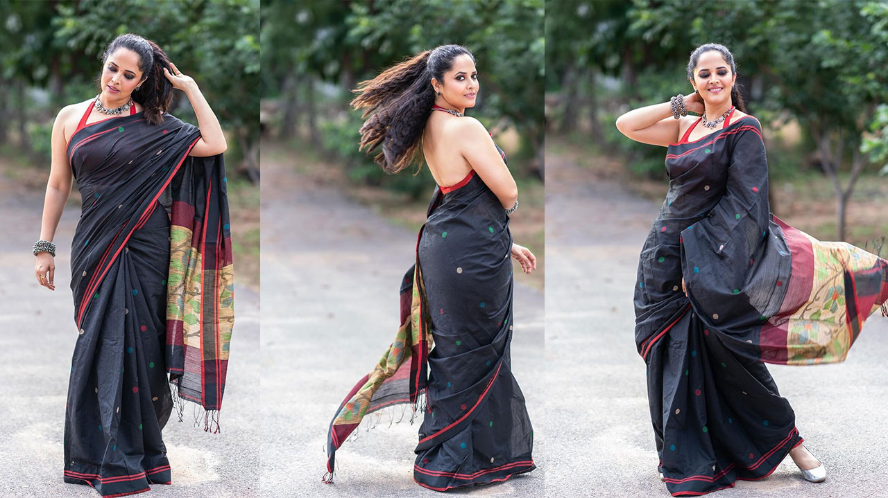 Anasuya Bharadwaj is wearing a draped garment with red borders and scattered dots, adorned with silver jewelry, outdoors among green foliage.