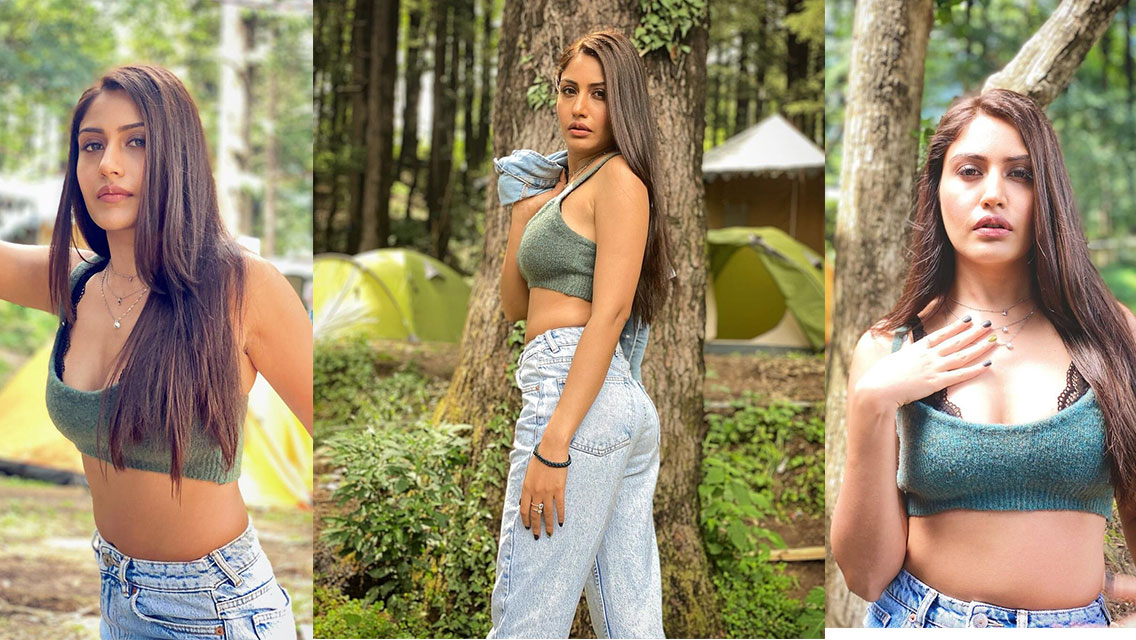 Surbhi Chandna stands in a wooded setting wearing a cropped knit top and ripped denim jeans, leaning against a large tree trunk.