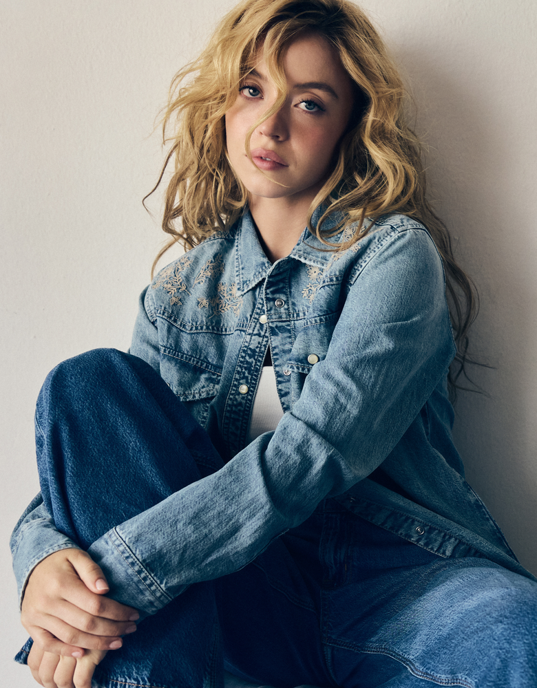 Sydney Sweeney in a three-quarter profile, draped in light wash blue denim against a neutral gray backdrop.