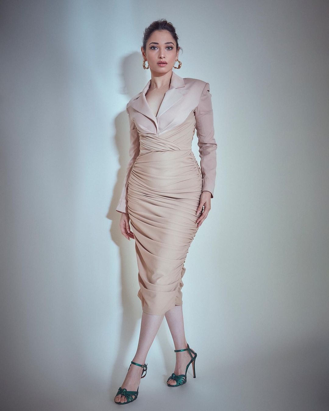 Tamannaah Bhatia wearing a flowing beige gown with a tailored jacket and gold earrings in a studio setting.