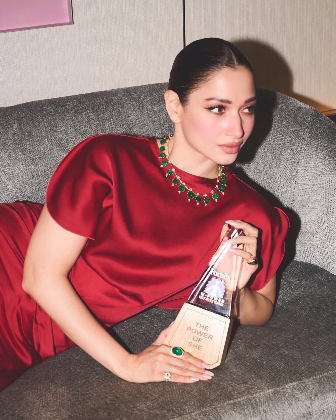Tamannaah Bhatia posing indoors in a deep red satin gown, seated on a sofa, surrounded by patterned rugs and artwork.