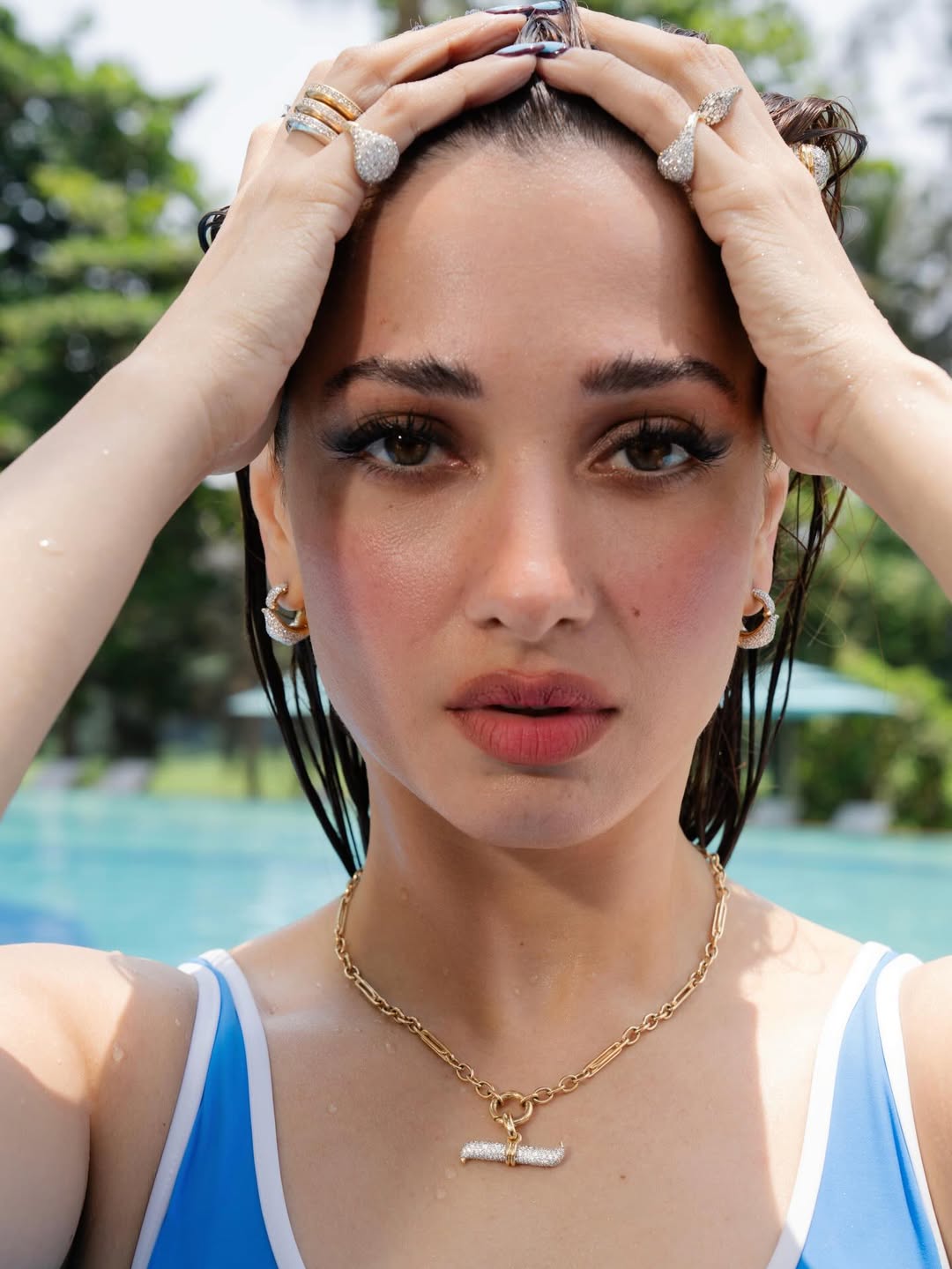 Tamannaah Bhatia reclining on a beige sofa wearing a white t-shirt and gold chain necklace.
