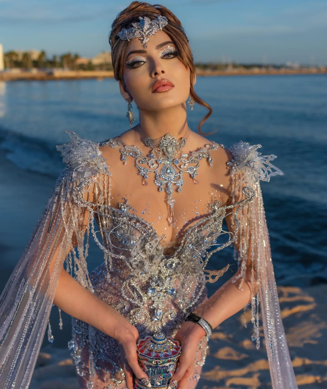 Urvashi Rautela posing outdoors holding an ornate blue and silver vase, wearing a heavily embellished gown by the ocean.