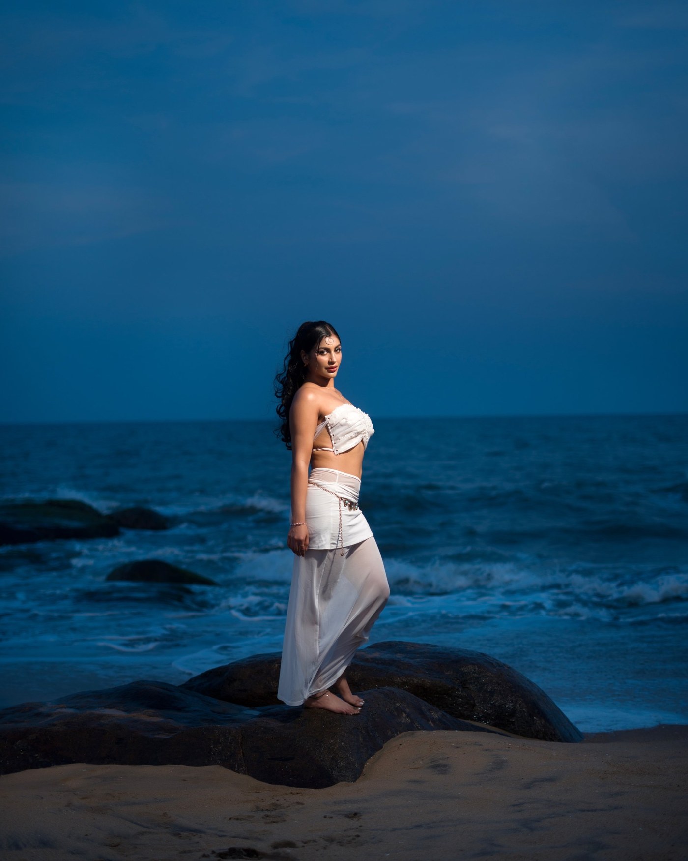 Yashika Aannand is captured serving glamorous elegance with a pearl-embellished white ensemble against an ocean backdrop.